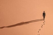 Eine Person läuft durch rötlichen, gewellten Wüstensand. Sie hinterläßt Spuren und wirft einen langen Schatten.
