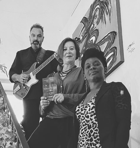 Schwarz-Weiß Foto: Auf einer Treppe stehen von hinten oben nach vorne Sazan Azodi mit Gitarre, Anette Schmitt mit dem Buch "Kleiner Bruder" und die Sängerin Dianka Kuamé.