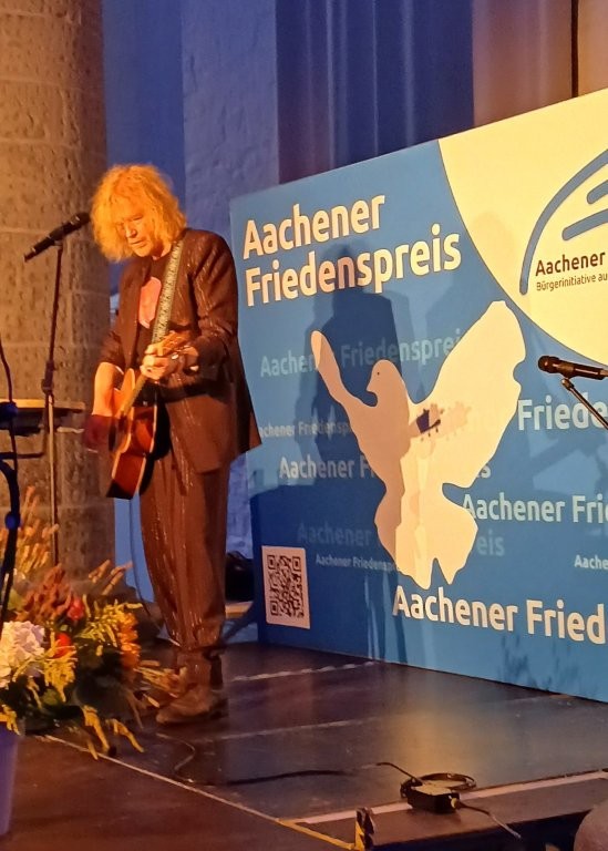 Eine Person spielt auf der Bühne Gitarre, im Hintergrund ist ein Banner des Aachener Friedenspreis.