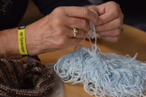 Hände mit einem Amnesty-Armband stricken hellblaue Wolle, die auf einem Tisch liegt.