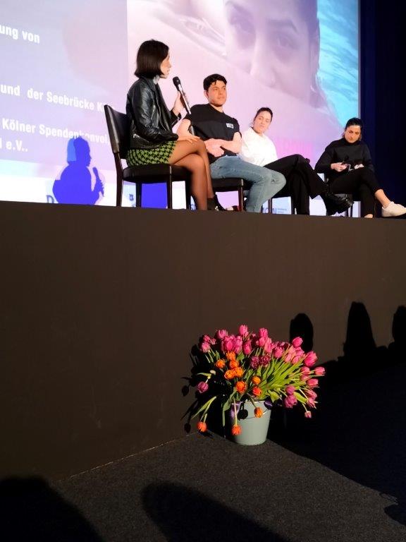 Charly Wai Feldman, Séan Binder und Sarah Mardini auf einer Bühne vor einer Kinoleinwand, vor der Bühne ein Eimer mit rosa Tulpen