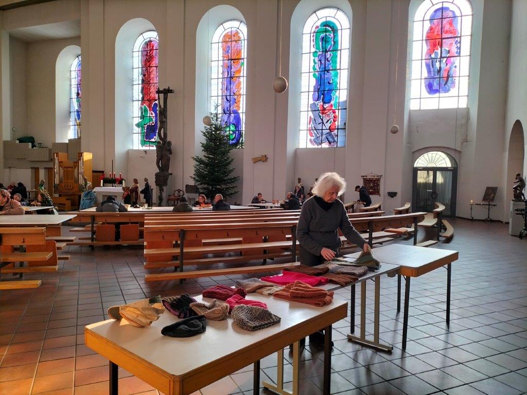 Während Menschen in der Kirche im Hintergrund an den Tischen sitzen, essen und sich unterhalten, werden vorne von einem Mitglied der Strickgruppe neue Strickwaren ausgelegt.