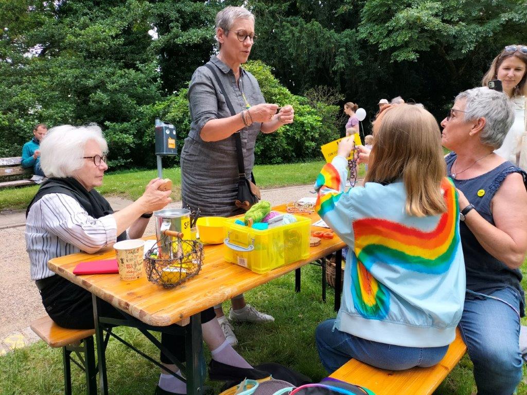 An einer Bierzeltgarnitur im Aachener Stadtpark zeigen Teilnehmer*innen des Strickcafés verschiedene Techniken. OB Sybille Keupen schaut vorbei.