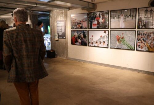 Fotoausstellung in einem Kellerraum mit Betonwänden. Im Hintergrund sind acht Fotos an der Wand zu sehen. Es sind einige Menschen im Raum verteilt.