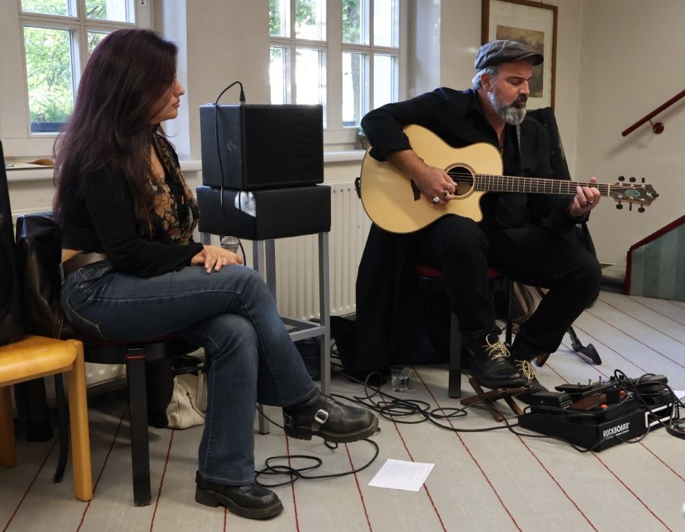 Ein Mann sitzt auf einem Stuhl und spiel Gitarre. Eine Frau sitzt daneben und hält ein Mikrofon.