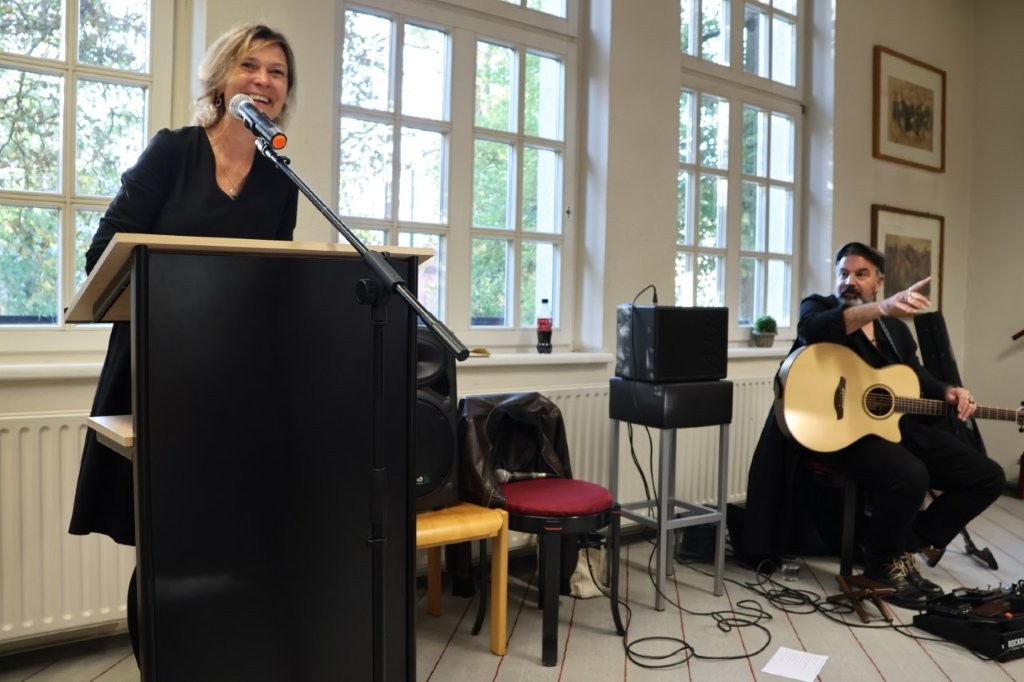 Eine Frau steht an einem Pult, im Hintergrund sitzt ein Mann mit einer Gitarre.