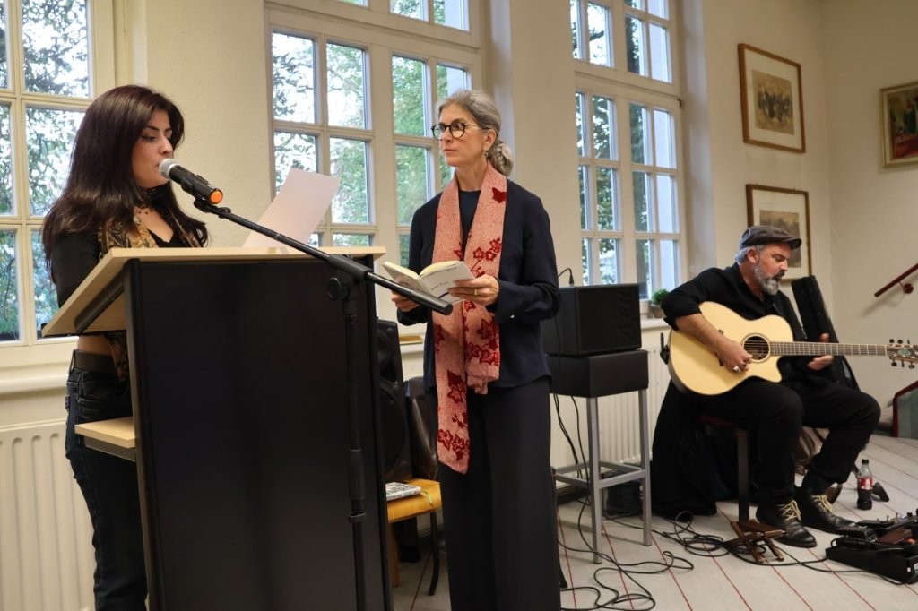 Eine Frau trägt an einem Pult etwas vor. Eine weitere Frau wartet mit einem Buch in der Hand. Im Hintergrund sitzt ein Mann mit einer Gitarre.