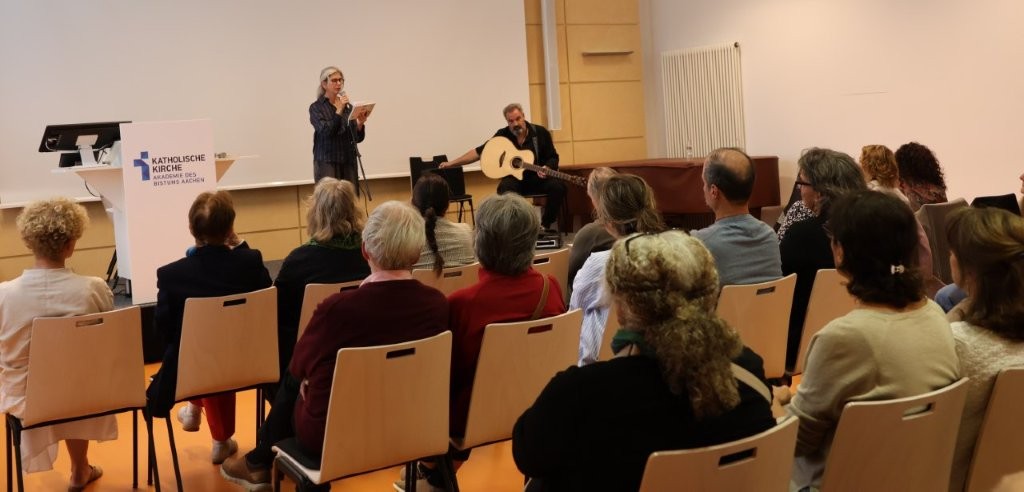 Blick über ein Publikum zu einer Bühne, eine Frau trägt einen Text vor, daneben sitzt ein Gitarrist.