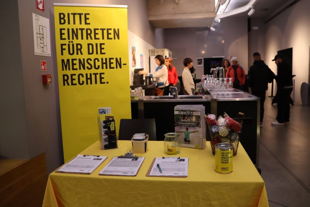 Amnesty Infostand mit gelber Tischdecke, Petitionen und Kerzen. Im Hintergrund eine Bar und ein Roll-Up: "Bitte eintreten für die Menschenrechte."