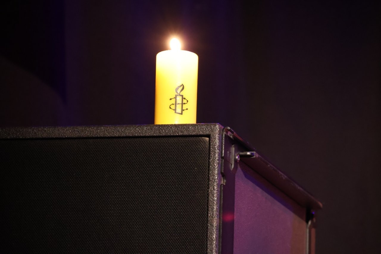 Eine gelbe Amnesty-Kerze brennt vor dunklen Hintergrund. Sie steht auf einer Lautsprecherbox.