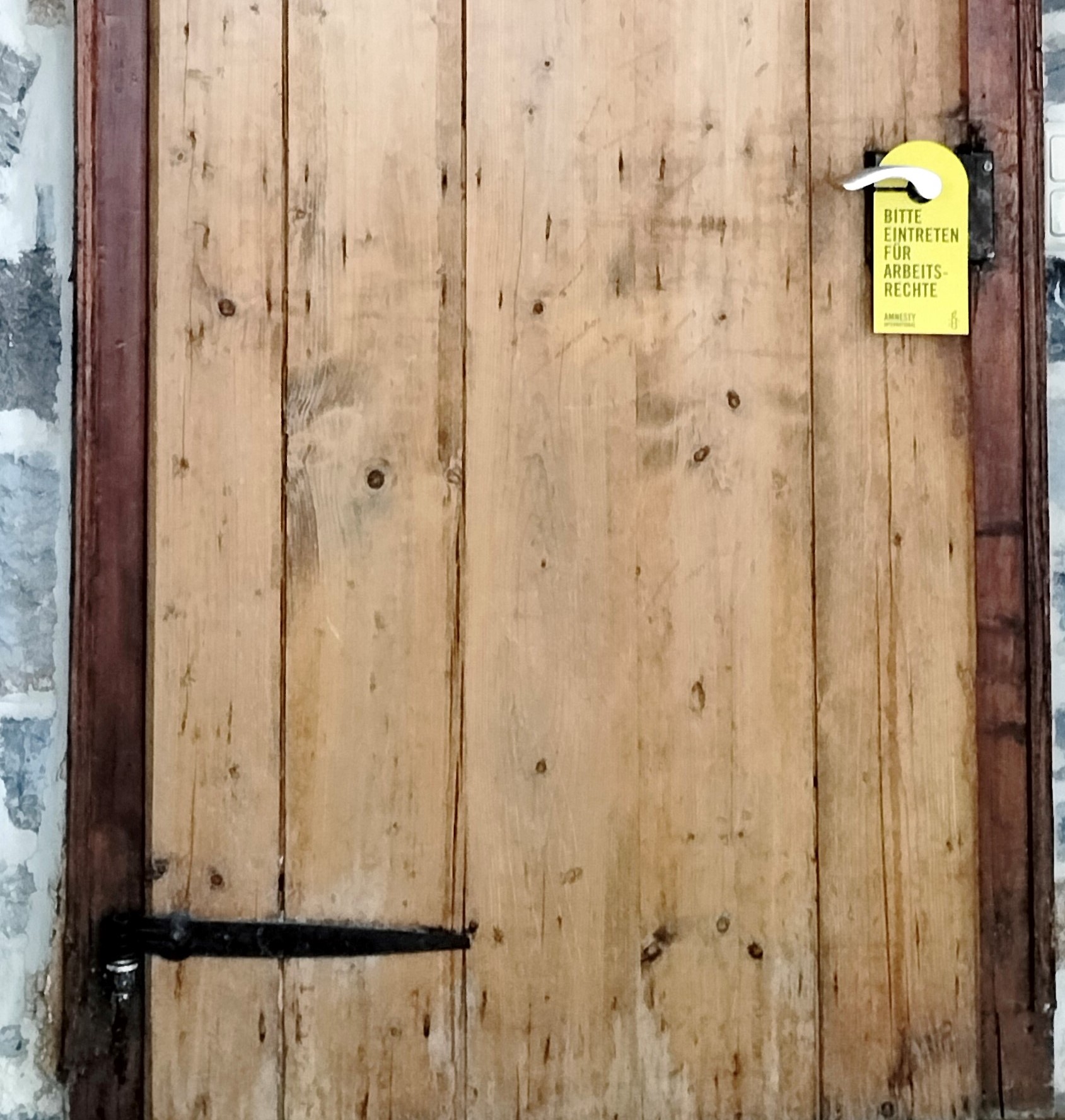 Tür aus alten Holzbrettern mit schwarz lackierten Holzbeschlägen. An der Klinke hängt ein gelbes Schild mit Aufschrift "Bitte eintreten für Arbeitsrechte".